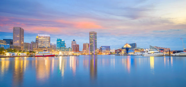 River by illuminated buildings against sky during sunset