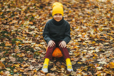 A boy a teen child in a bright yellow hat sits on a pumpkin in an autumn forest in nature outdoors