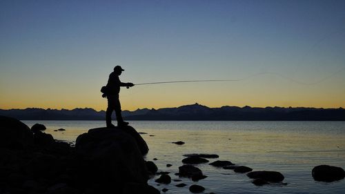 Silhouette man standing on rock by sea against clear sky during sunset