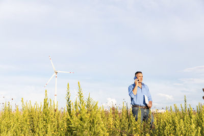 Male engineer talking through mobile phone and document while standing against wind turbines on field