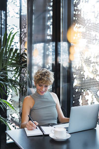 Freelancer with laptop on table writing in diary at coffee shop