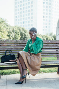 Serious young black female executive with short dyed hair in trendy outfit talking smartphone while sitting on wooden bench in park