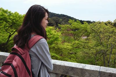 Side view of young woman standing by railing against trees