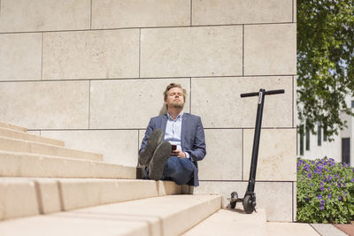 Businessman with e-scooter and smartphone sitting on steps relaxing