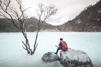 Man sitting on rock by lake against mountain