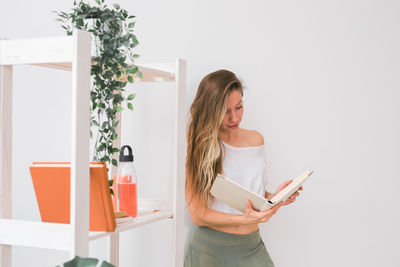 Woman reading book while standing against wall at home