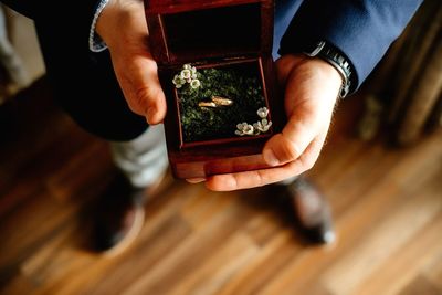 Low section of man holding wedding rings while standing on hardwood floor