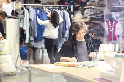 Man working on sewing machine while volunteer searching for clothes in rack at workshop