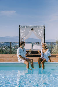 Couple sitting by swimming pool against sky