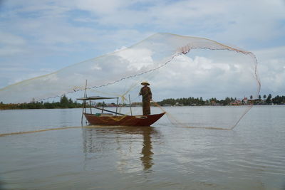 Fisherman throwing net while standing in boat on sea against sky