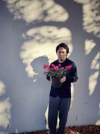 Young asian man holding red flowering cyclamen potted plant against shadow patterned wall.