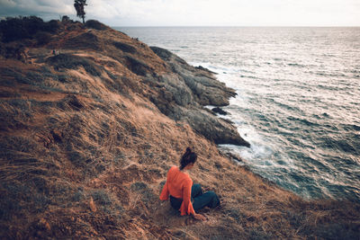 Rear view of man sitting on rock by sea against sky