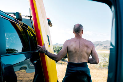 Back view of active middle aged shirtless man taking paddle board from automobile parked on lake shore in hilly nature while preparing for water sport practice in summer day