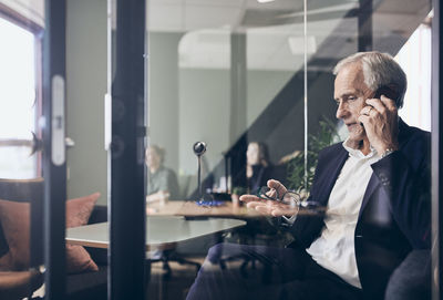 Mature male manager talking on mobile phone seen through glass wall at creative office