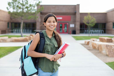 Portrait of young girl in a backpack holding books standing in front of a school entrance 