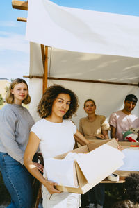 Portrait of young female entrepreneur holding cardboard box while standing with colleagues near tent at community center