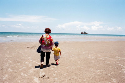Rear view of grandmother and grandson walking at beach against sky