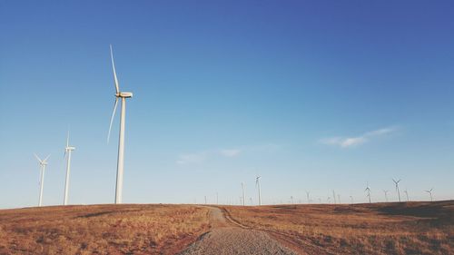 Dirt road on field by windmills against sky