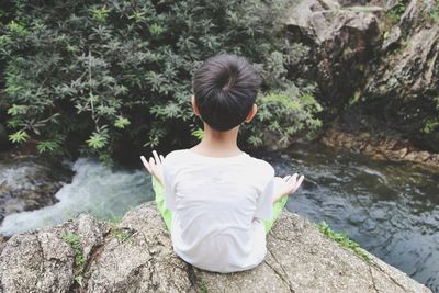 Rear view of boy sitting on rock over stream at forest