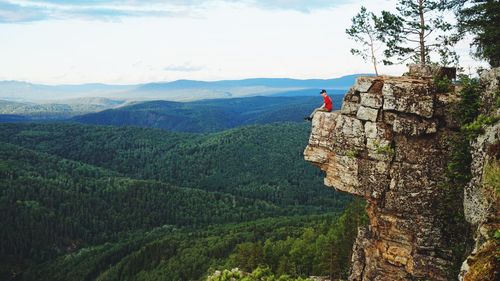 Man sitting on cliff over landscape against sky