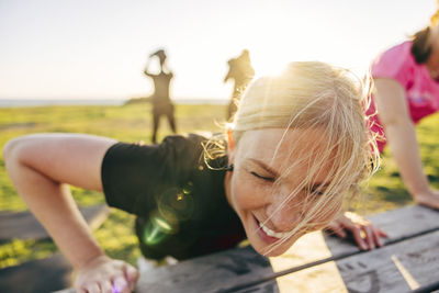 Smiling blond woman doing push-ups on bench during group training at beach