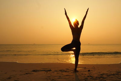 Full length of woman practicing yoga on shore at beach against sky during sunset