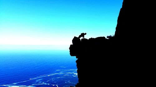 Silhouette man exercising on cliff by sea against clear blue sky