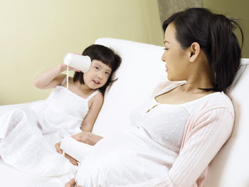 Girl listening pregnant mother abdomen with tin can phone on sofa