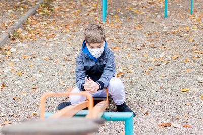 Sad kid wearing face mask while sitting on seesaw at empty playground during coronavirus pandemic.