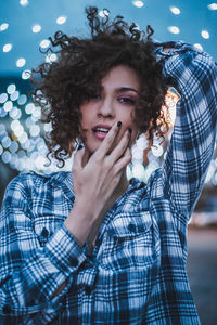 Portrait of young woman with hand in hair standing against illuminated string lights