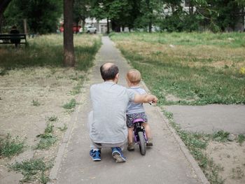 Rear view of father teaching son to ride bicycle on footpath