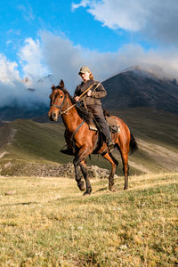 Side view of a cowgirl shepherd riding on a horse on a mountain valley against sky with clouds