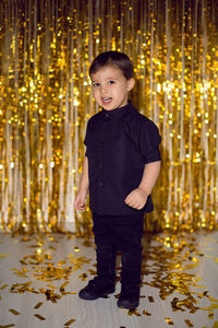 Boy in black clothes at christmas standing in a room on a background of gold tinsel on the wall