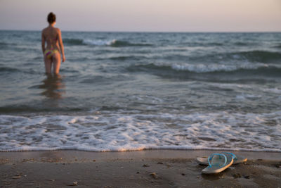 Rear view of young woman wearing bikini while standing in sea against sky during sunset