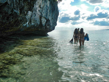 Rear view of friends walking in sea by rock formation against cloudy sky