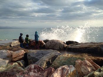 Rear view of friends standing on rocky shore against cloudy sky