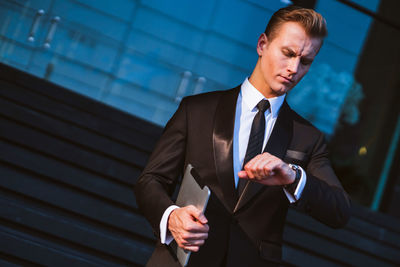 Handsome businessman with digital tablet checking time while standing against building in city