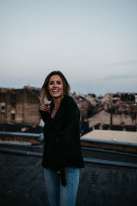 Portrait of smiling young woman standing against cityscape during sunset