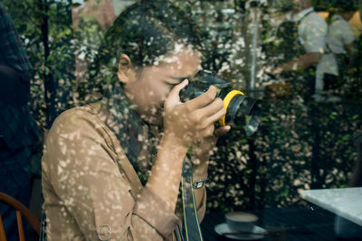 Side view of woman photographing while sitting at cafe seen through glass window