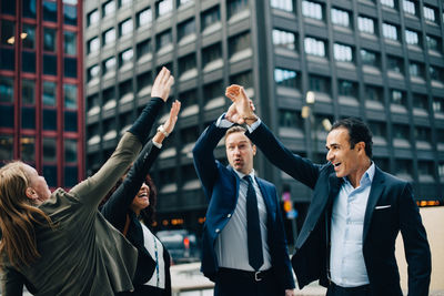 Smiling multi-ethnic business people standing with hands raised against building in city