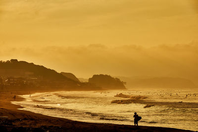 Silhouette man walking with surfboard at beach against orange sky during sunrise