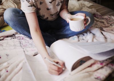 Midsection of woman holding coffee studying while sitting on bed at home