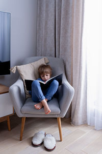 Cute preschool blonde kid reading book sitting on a cozy chair in light nordic style living room