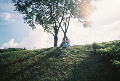 Woman hugging tree while sitting on grassy field against sky