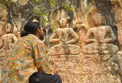 Rear view of a long haired young guy sitting in front of gyalwa ringna 5 dhyani buddha rock statue.