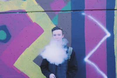 Portrait of young man smoking while standing against graffiti wall
