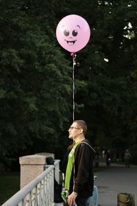 Side view of man standing with balloon by railing against trees