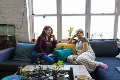Smiling female friends sitting together on sofa against window at home