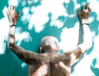 Rear view of shirtless man with arms raised leaning on wall