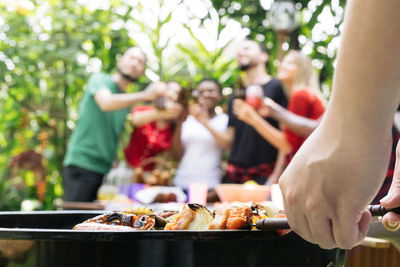 Cropped hand preparing food on barbecue grill with friends in background